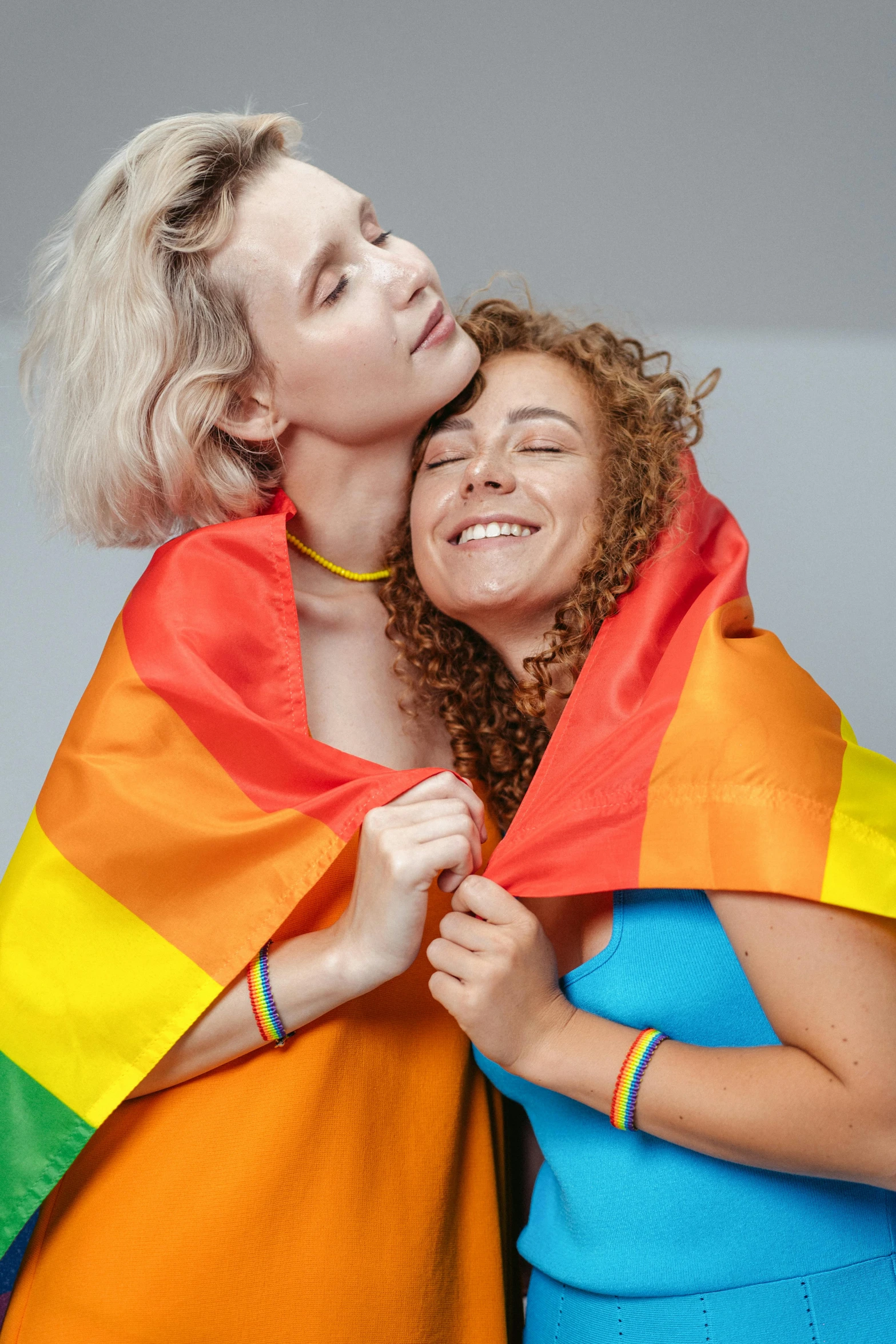 two women wrapped in a rainbow colored blanket, a photo, shutterstock, antipodeans, short curly blonde haired girl, lgbt flag, curly haired, two men hugging