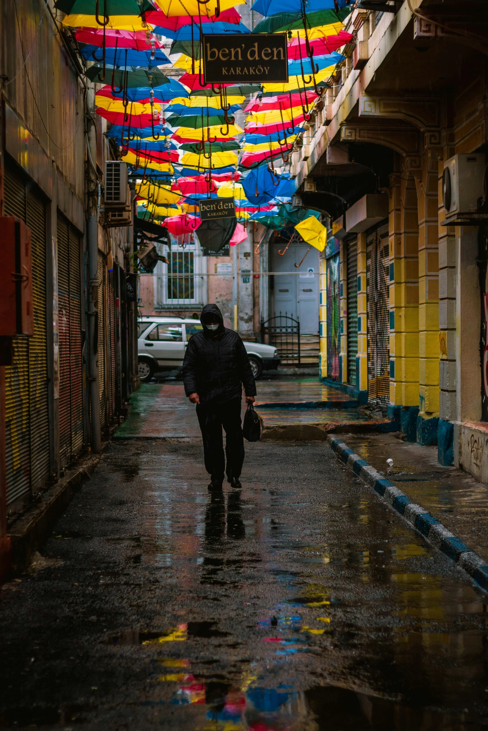 a person walking down a wet street under umbrellas, by Ibrahim Kodra, unsplash contest winner, street art, masked person in corner, turkey, quiet, multiple colors