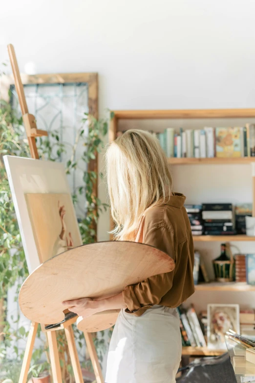 a woman standing in front of a painting on a easel, trending on unsplash, sitting on a mocha-colored table, holding a shield, botanicals, architect studio