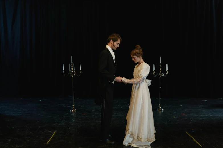 a man and woman standing next to each other on a stage, by Alice Mason, unsplash, renaissance, in a long white dress, black victorian dress, candle lighting, cinema still