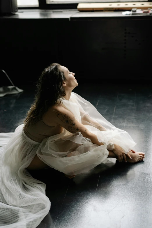 a woman in a white dress sitting on the floor, by Elizabeth Polunin, unsplash, light and space, tattooed, video still, studio picture, looking off to the side