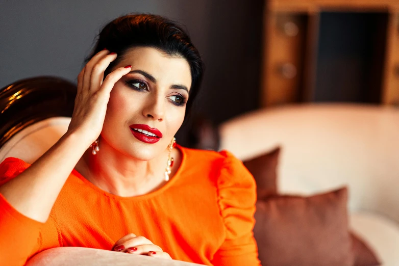 a woman in an orange dress sitting on a couch, a portrait, instagram, hurufiyya, shohreh aghdashloo, taken with sony alpha 9, lipstick, middle eastern