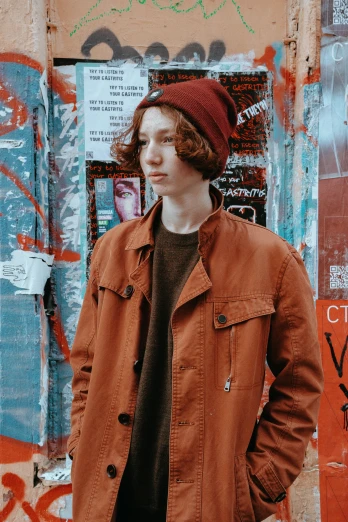 a man standing in front of a wall covered in graffiti, an album cover, trending on pexels, realism, brown reddish hair, beanie, very very very pale skin, orange jacket