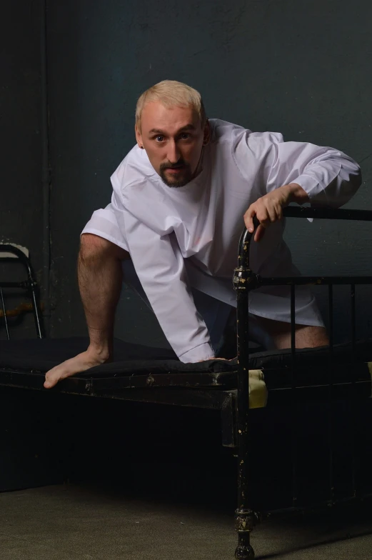 a man leaning over a bed in a dark room, an album cover, inspired by Dmitry Levitzky, wearing a white hospital gown, richard garriott, shaved head, photo taken in 2018