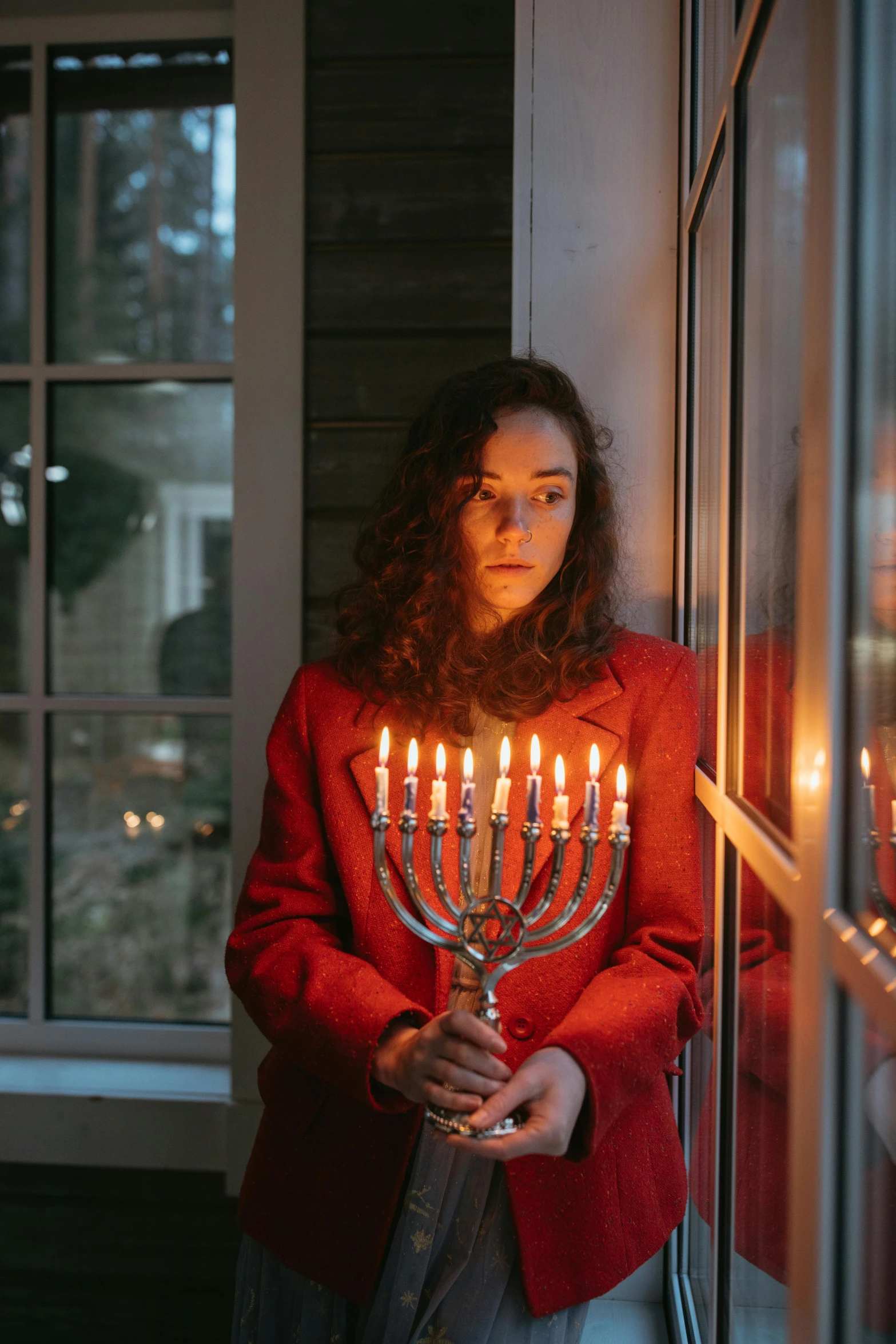 a woman holding a lit menorah in front of a window, by Harriet Zeitlin, pexels, wearing red jacket, instagram post, teenager, (1 as december