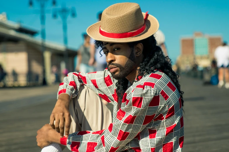 a man sitting on a boardwalk wearing a hat, an album cover, by Pamela Ascherson, pexels, renaissance, brown skin man egyptian prince, red suit, square, 15081959 21121991 01012000 4k