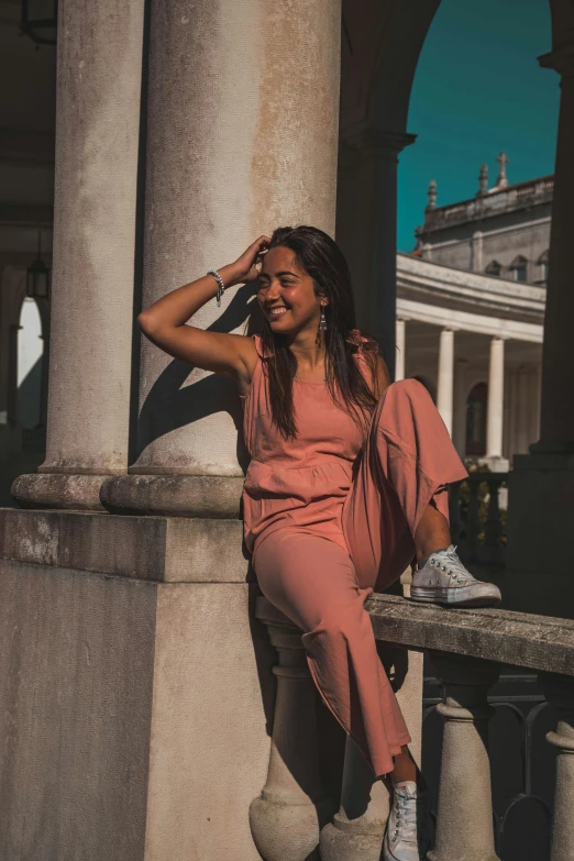 a woman sitting on a wall talking on a cell phone, by Emma Andijewska, pexels contest winner, renaissance, jumpsuit, satisfied pose, with soft pink colors, tanned