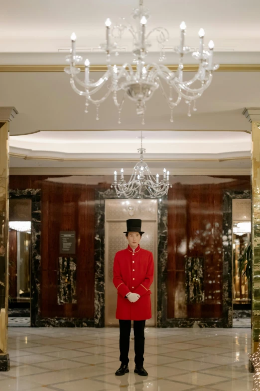 a man in a red coat standing in front of a chandelier, hotel, embroidered uniform guard, 8ft tall, staring