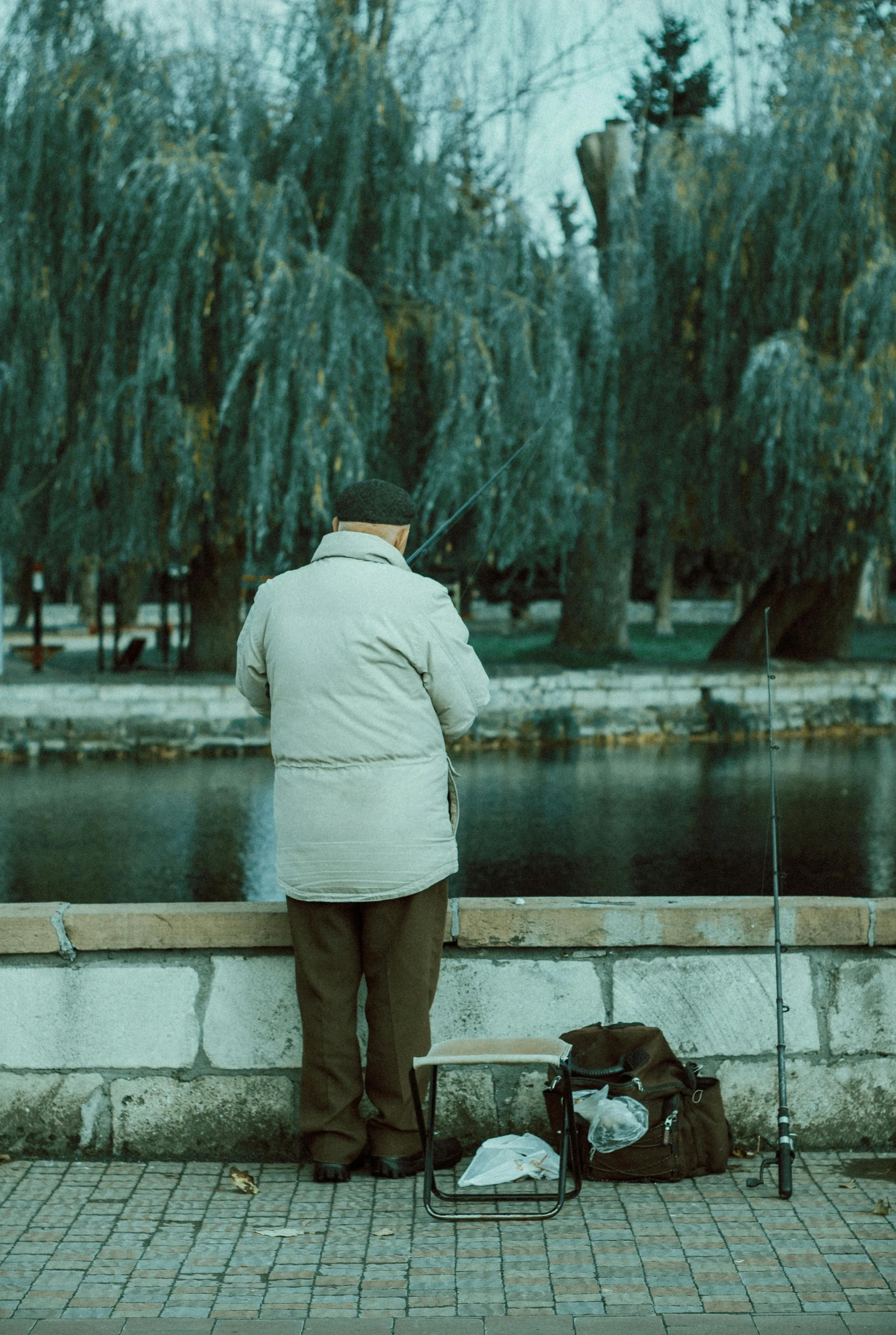 a man sitting on a bench next to a body of water, fishing, standing next to water, looking old, at a park
