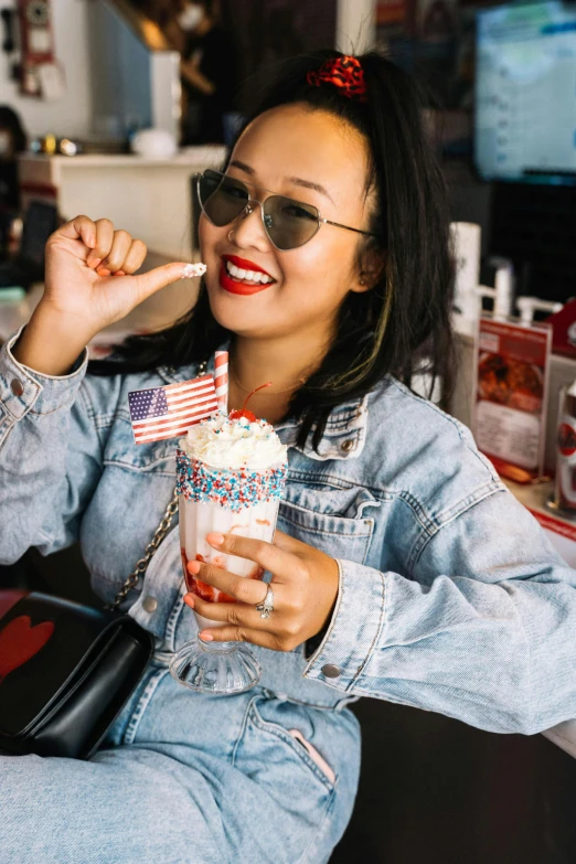 a woman sitting at a table with a drink in her hand, a portrait, by Julia Pishtar, trending on unsplash, american flags, ice cream on the side, wearing denim, asian descent