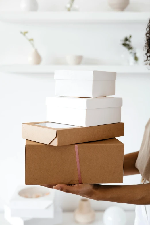 a woman holding a stack of boxes in her hands, by Eden Box, browns and whites, paper craft, manuka, holiday season