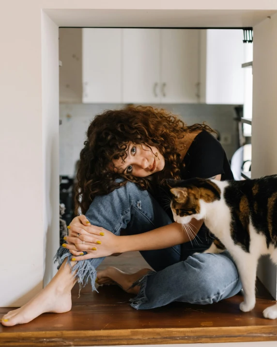 a woman sitting on a counter petting a cat, by Julia Pishtar, pexels contest winner, brown curly hair, sitting on the floor, queer woman, lovingly looking at camera