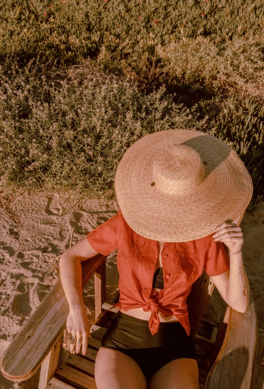 a woman sitting in a chair with a hat on, pexels contest winner, sunfaded, red hue, covered in sand, lush surroundings