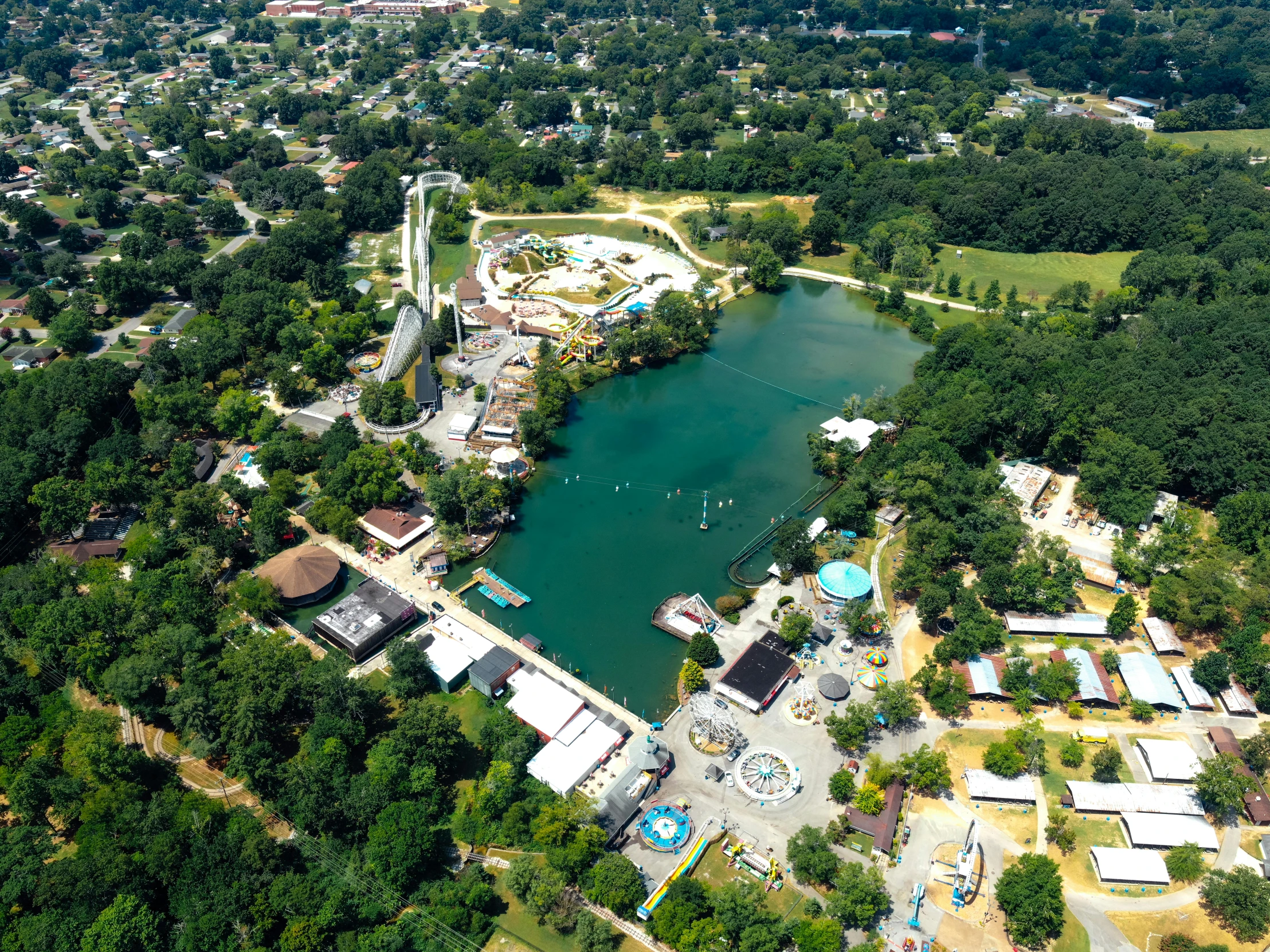 a large body of water surrounded by trees, amusement park buildings, bentonville arkansas, looking down on the camera, splash image