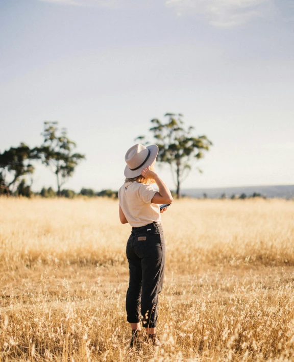 a woman in a hat standing in a field, inspired by Russell Drysdale, trending on unsplash, happening, plain uniform sky at the back, on a hot australian day, wearing farm clothes, hands on hips