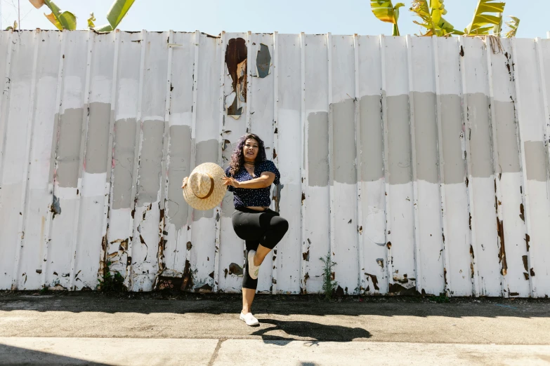 a woman standing in front of a white fence holding a hat, inspired by Gina Pellón, pexels contest winner, street art, in a jumping float pose, in front of a garage, 15081959 21121991 01012000 4k, hispanic