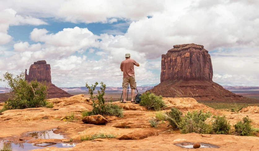 a man standing on top of a rock next to a body of water, by Lee Loughridge, pexels contest winner, plein air, monument valley, background image, conde nast traveler photo, looking at the ground