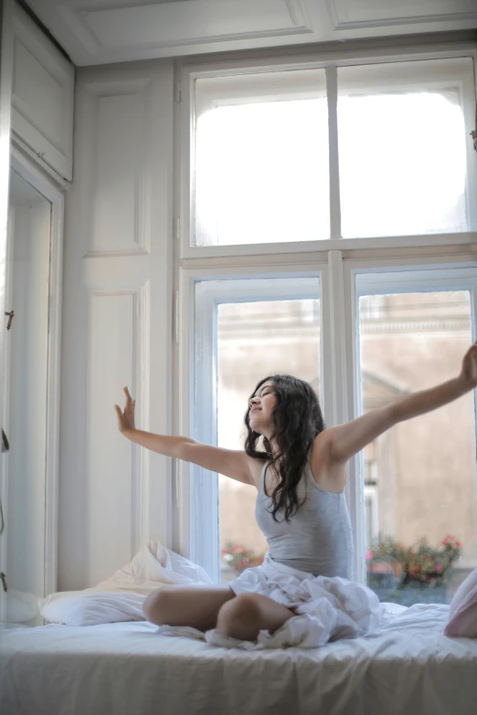 a woman sitting on top of a bed next to a window, pexels contest winner, arabesque, with arms up, square, :: morning, brunette fairy woman stretching