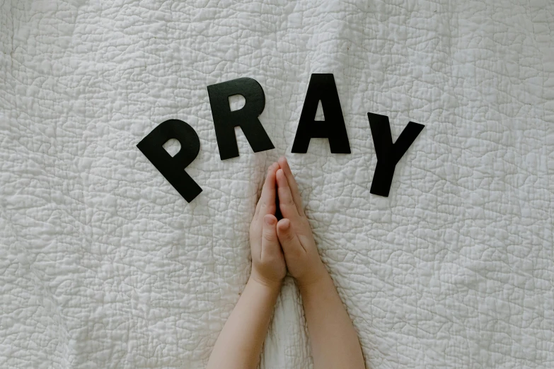 a person laying on a bed with the word pray spelled in black letters, by Carey Morris, pexels, little boy wearing nun outfit, hands pressed together in bow, 1 6 x 1 6, play