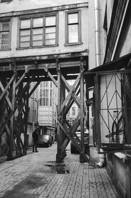 a black and white photo of a street, wooden supports, location [ chicago ( alley ) ], metallic bridge, mechanical structure