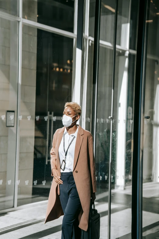 a woman wearing a face mask standing in front of a building, trending on pexels, in a hotel hallway, light brown coat, walking to work, worksafe. instagram photo