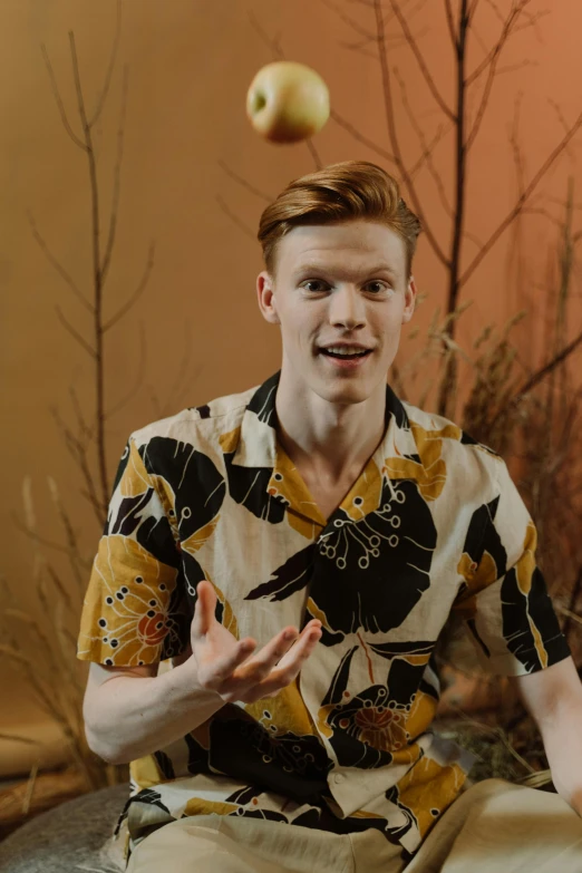 a man sitting on a rock with an apple in the air, an album cover, by Jacob Toorenvliet, trending on pexels, renaissance, ginger hair with freckles, wearing yellow floral blouse, handsome man, video still