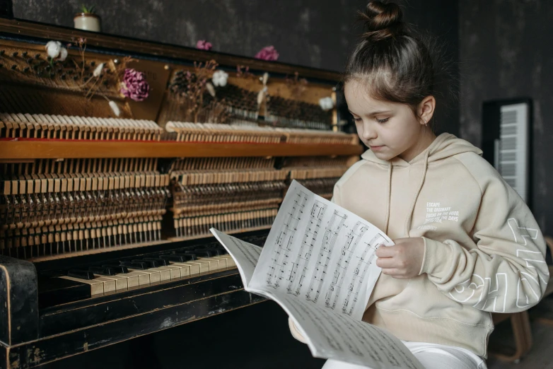 a young girl reading a sheet of music in front of a piano, pexels contest winner, danube school, sydney hanson, 15081959 21121991 01012000 4k, girl wearing hoodie, ornately dressed
