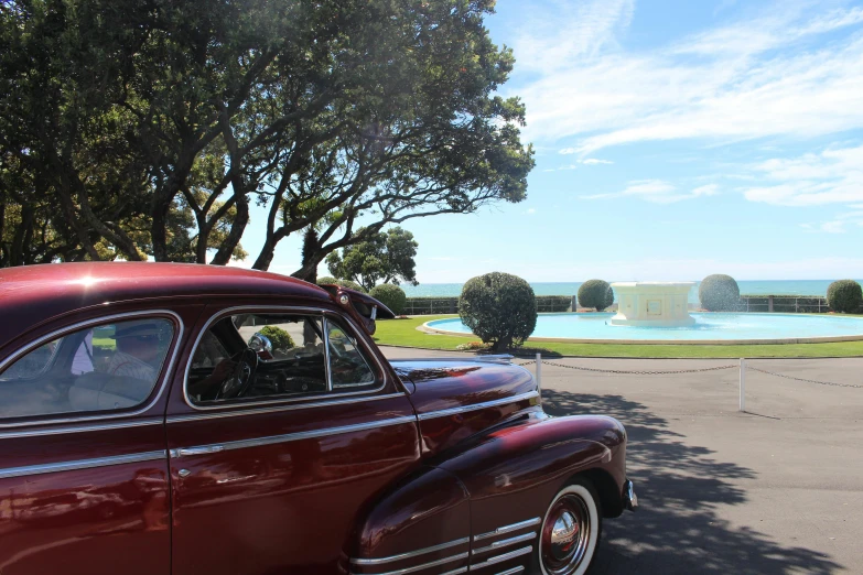 a classic car parked in front of a fountain, views to the ocean, clear blue skies, te pae, payne's grey and venetian red