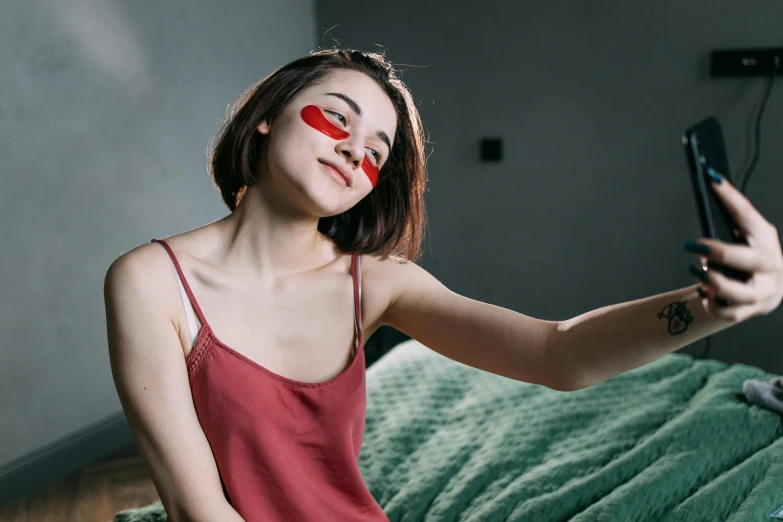 a woman sitting on a bed taking a selfie, trending on pexels, red paint strip across eyes, with round cheeks, avatar image, playful pose