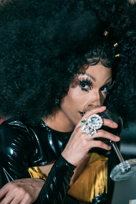 a woman sitting at a table with a glass of wine, an album cover, trending on pexels, renaissance, ru paul\'s drag race, drinking champagne, performing on stage, black curly hair