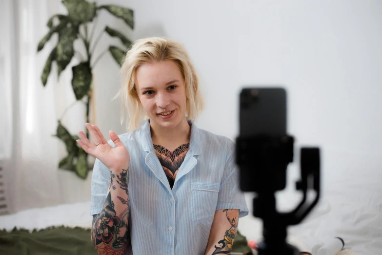 a woman sitting on top of a bed next to a camera, a tattoo, trending on pexels, video art, avatar with a blond hair, webcam footage, giving a thumbs up, smartphone photo