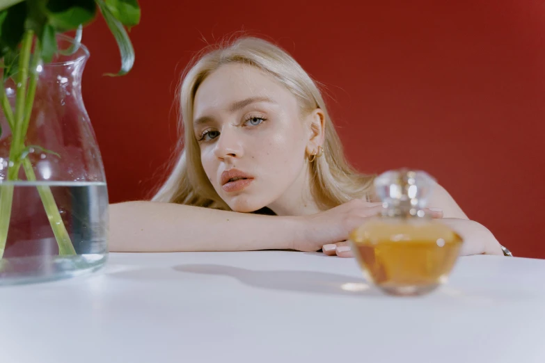 a woman sitting at a table next to a vase of flowers, inspired by Elsa Bleda, trending on pexels, photorealism, floating in perfume, blonde swedish woman, sydney sweeney, smooth red skin
