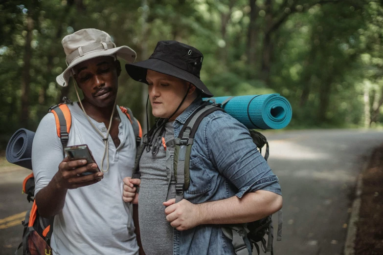 a couple of men standing next to each other on a road, trending on pexels, renaissance, carrying survival gear, looking at his phone, wearing a travel hat, black man