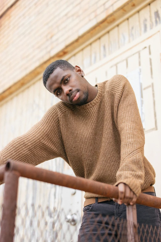 a man leaning on a rail in front of a building, an album cover, inspired by Theo Constanté, trending on pexels, realism, he is wearing a brown sweater, light-brown skin, striking a pose, corduroy