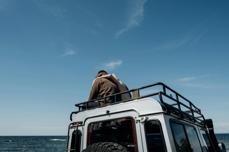 a man standing on top of a vehicle next to the ocean, unsplash, private press, land rover defender, clear blue skies, looking her shoulder, man sitting facing away