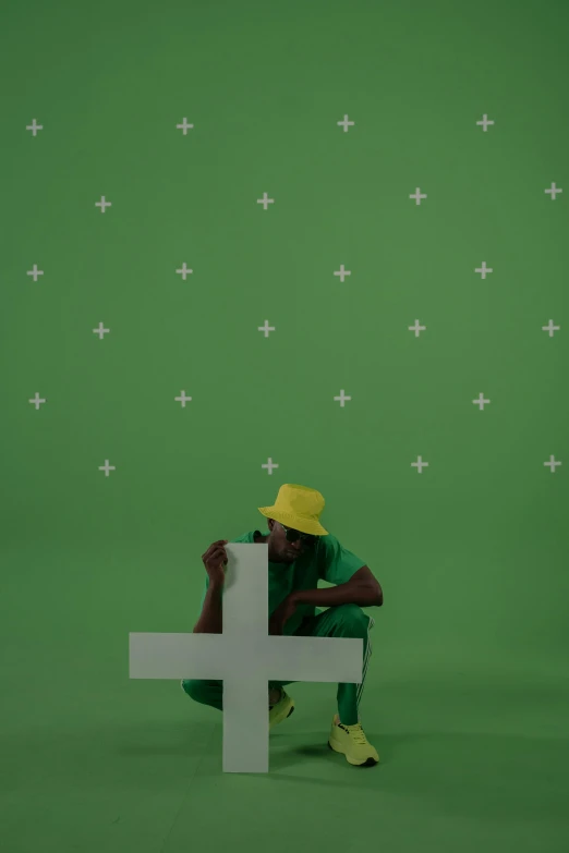 a man sitting in front of a cross on a green screen, an album cover, by artist, conceptual art, character with a hat, acids, ( ( dark skin ) ), performing a music video