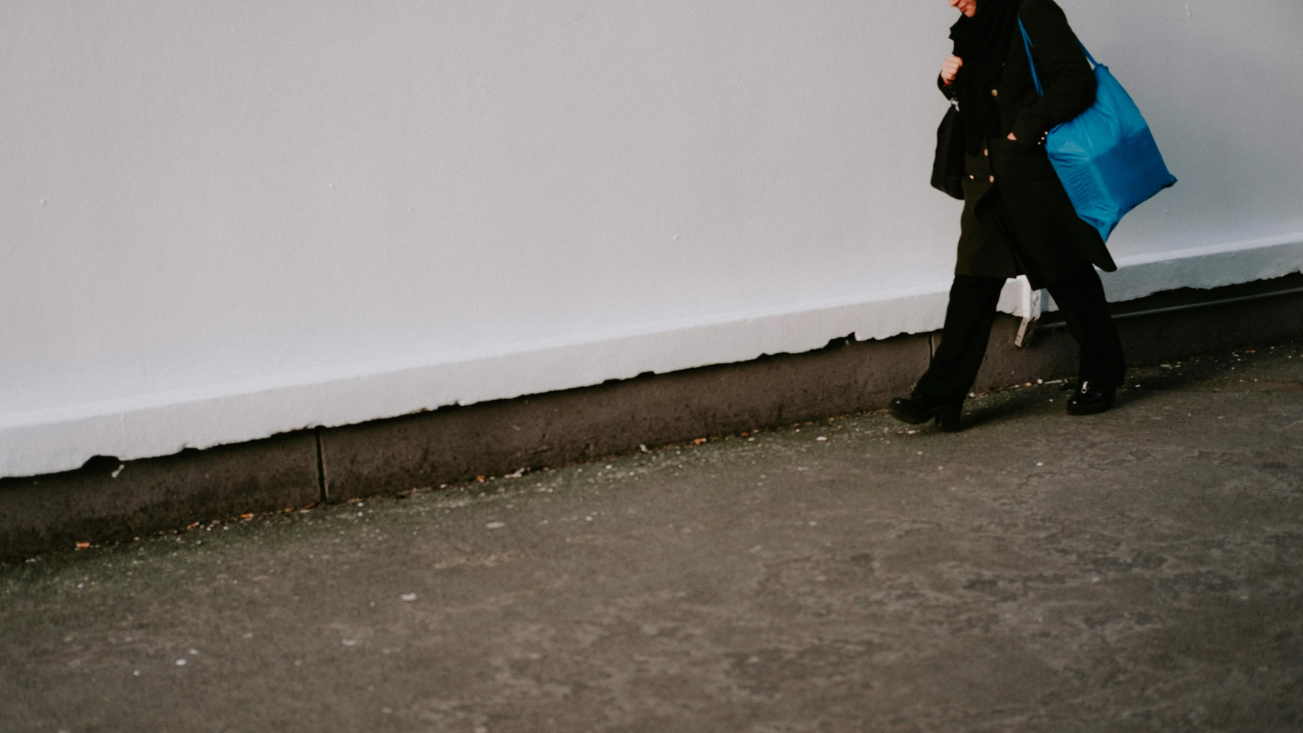 a woman walking down a sidewalk carrying a blue bag, a photo, trending on pexels, postminimalism, wall corner, wearing black boots, white wall complex, leaning on the wall