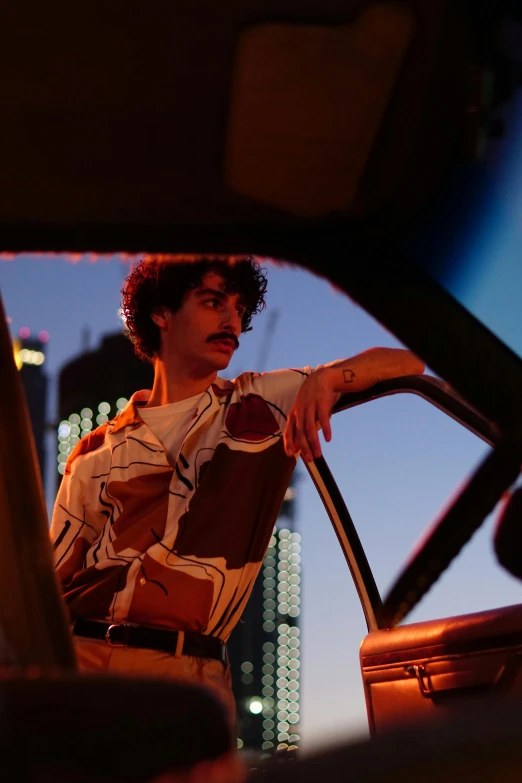 a man sitting in the driver's seat of a car, an album cover, inspired by Nan Goldin, trending on pexels, an arab standing watching over, unibrow, city view, model posing
