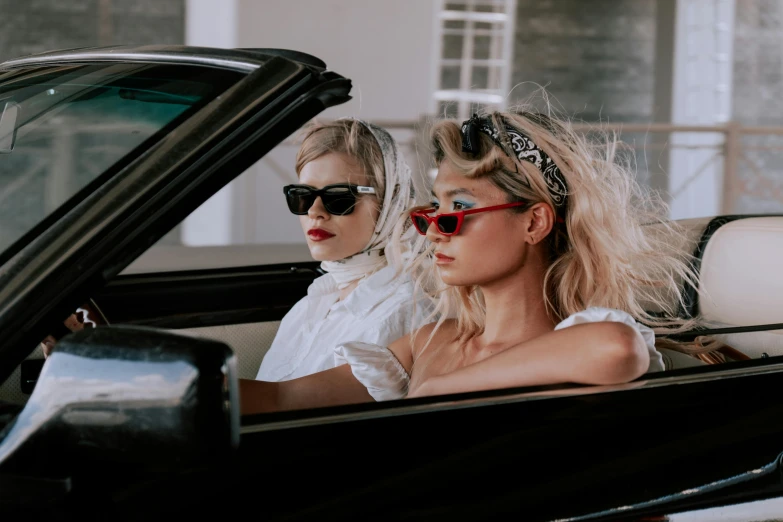 two women sitting in the driver's seat of a convertible car, an album cover, by Emma Andijewska, trending on pexels, blonde, wearing victorian goggles, at a fashion shoot, charli bowater and artgeem