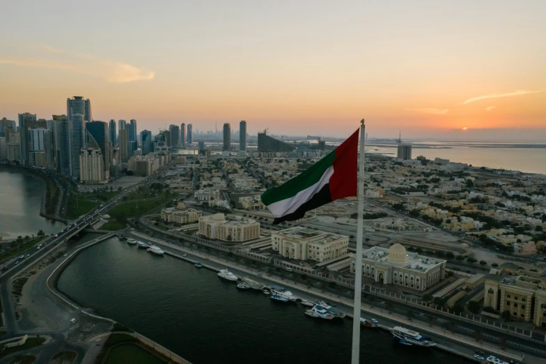 a flag flying on top of a tall building next to a body of water, by Julia Pishtar, pexels contest winner, hurufiyya, arab ameera al taweel, skyline view from a rooftop, with a sunset, slide show