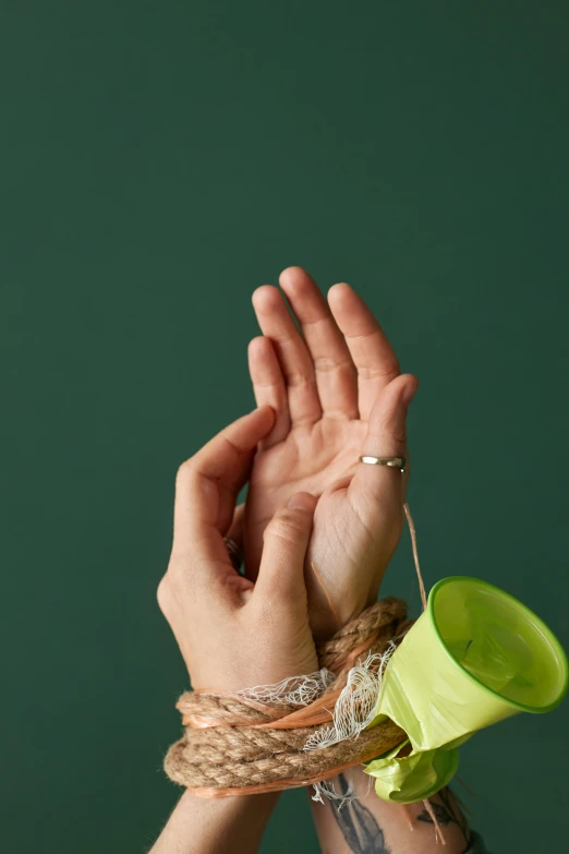 a woman's hands tied up with rope and a green cup, an album cover, inspired by Elsa Bleda, trending on pexels, plasticien, hand gesture, plain background, open palm, crafts