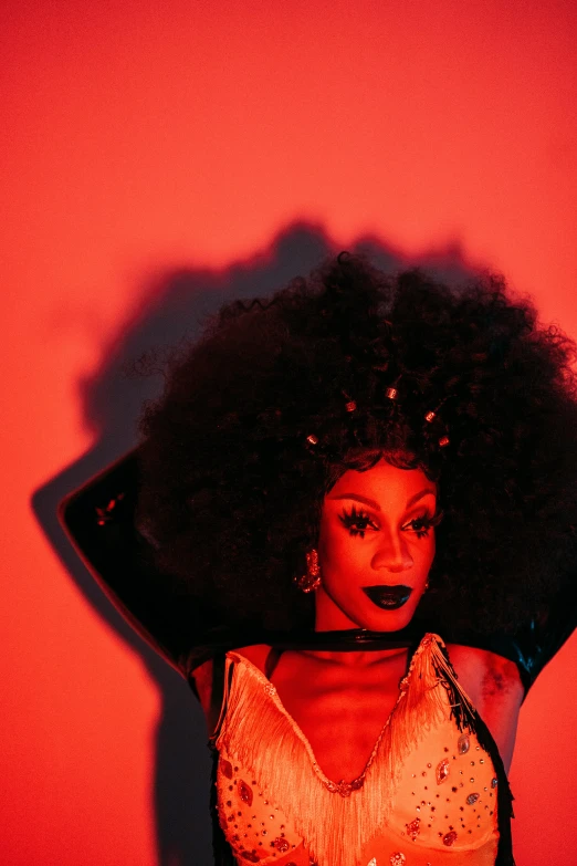 a woman standing in front of a red wall, an album cover, afrofuturism, drag queen, shot at dark with studio lights, wavy hair spread out, portrait of hisoka hunter hunter