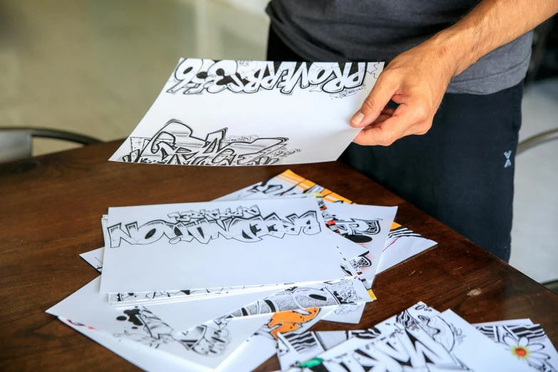 a man holding a piece of paper with drawings on it, by Tom Bonson, unsplash, letterism, graffiti letters, character sheets on table, many copies of them, comic art style