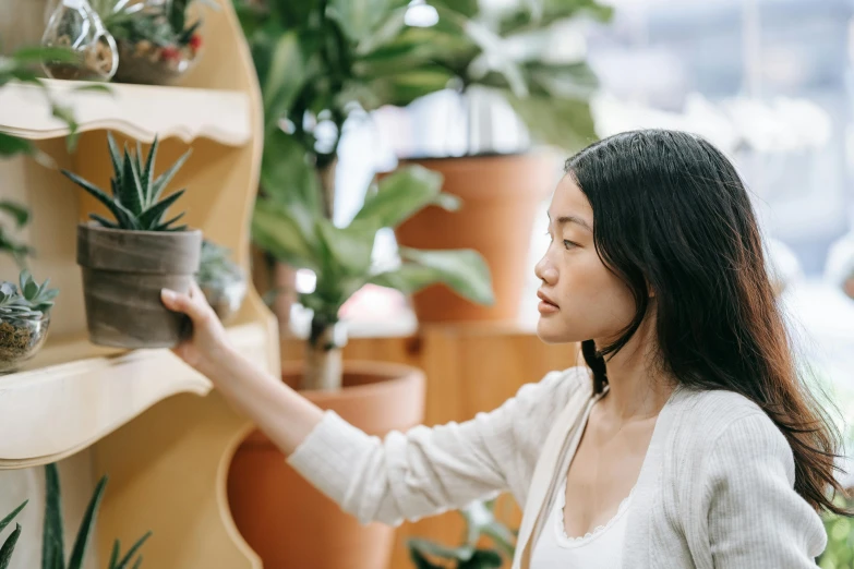 a woman standing in front of a shelf filled with potted plants, trending on pexels, arts and crafts movement, pondering, gemma chen, inspect in inventory image, close up shot from the side