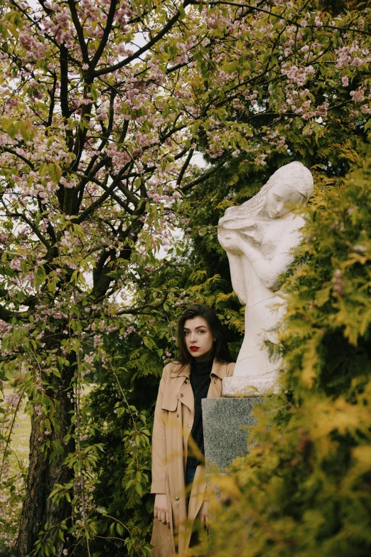 a woman standing next to a statue in a park, inspired by Elsa Bleda, unsplash, art nouveau, blossoms, dua lipa, woman is in a trenchcoat, still from a music video