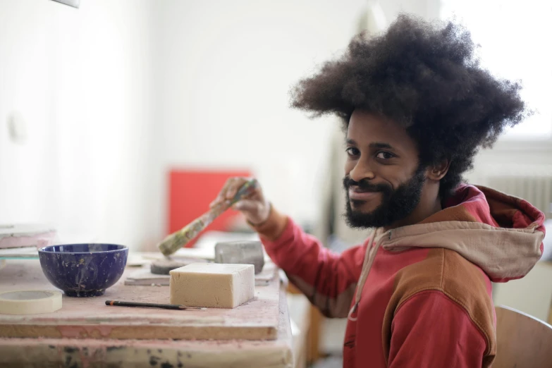 a man sitting at a table with a brush in his hand, a portrait, inspired by Afewerk Tekle, pexels contest winner, arbeitsrat für kunst, looking happy, with afro, ceramics, art student