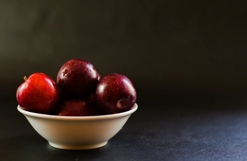 a bowl filled with red apples sitting on top of a table, in front of a black background, background image, plum color scheme, thumbnail