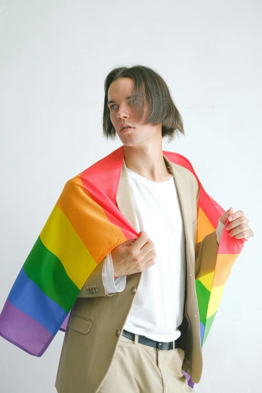 a man standing in front of a white wall holding a rainbow flag, an album cover, unsplash, bauhaus, wearing a brown cape, joe keery, open jacket, attractive androgynous humanoid