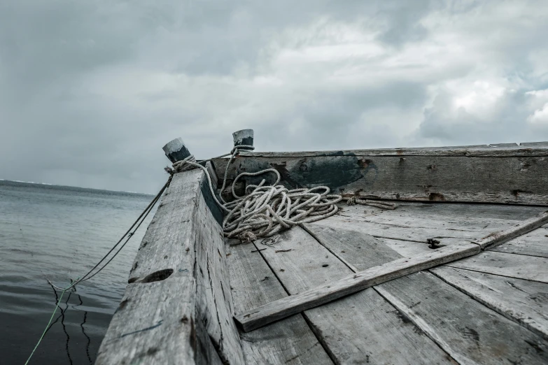 a wooden boat sitting on top of a body of water, by Sebastian Spreng, pexels contest winner, overcast gray skies, rope, damaged, looking from side