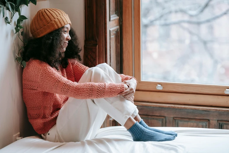 a woman sitting on top of a bed next to a window, trending on pexels, knitted hat, happily tired, coloured, seasonal